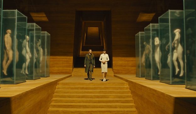 Ryan Gosling and Sylvia Hoeks walk past displays of replicants in Blade Runner 2049